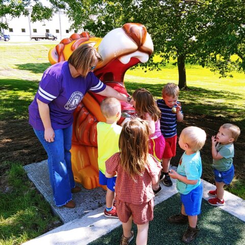 A woman dressed in jeans and a purple T-shirt with a Lions Club logo on it assists a group of small children surrounding a cartoonish lion statue with a drinking fountain in its mouth.