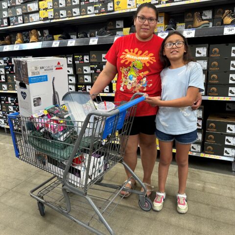 A woman in a red top and blue shorts has her arm around a pre-teen girl in a white t-shirt and denim shorts posing beside a shopping cart in the middle of a super center.