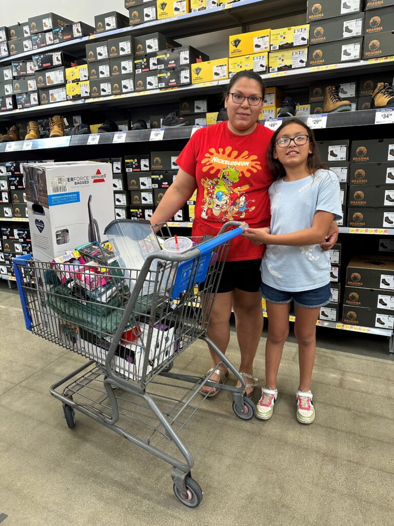 A woman in a red top and blue shorts has her arm around a pre-teen girl in a white t-shirt and denim shorts posing beside a shopping cart in the middle of a super center.